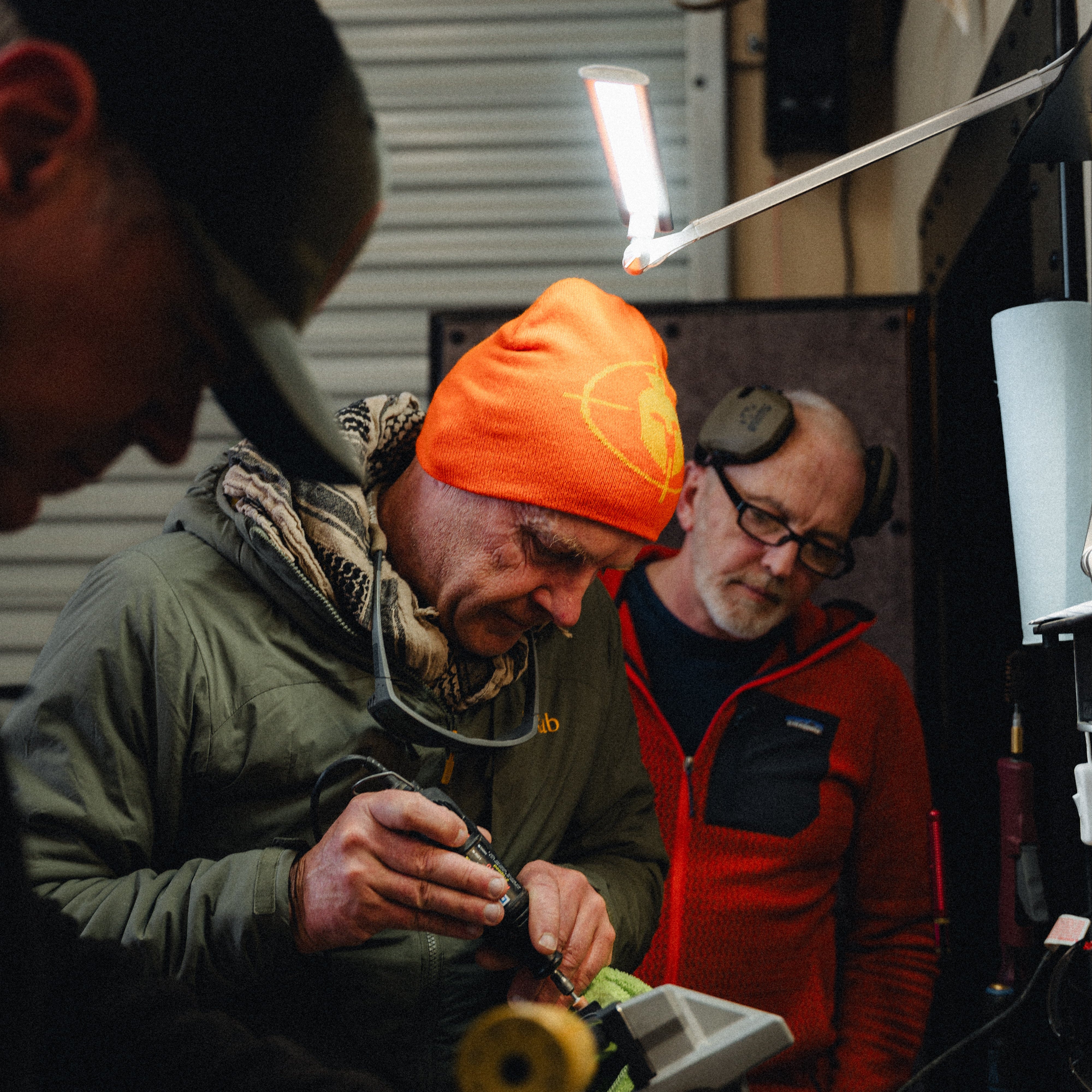 Two men working on a project with tools, one wearing an orange beanie and the other in a red jacket.