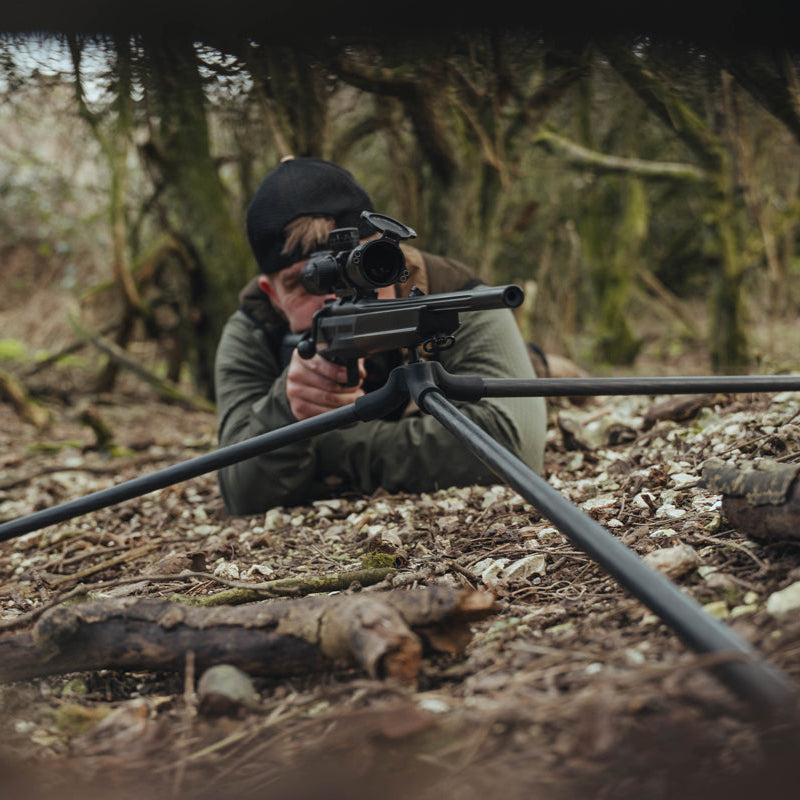 Person in a forest setting with a rifle, wearing a green jacket and black backwards cap.