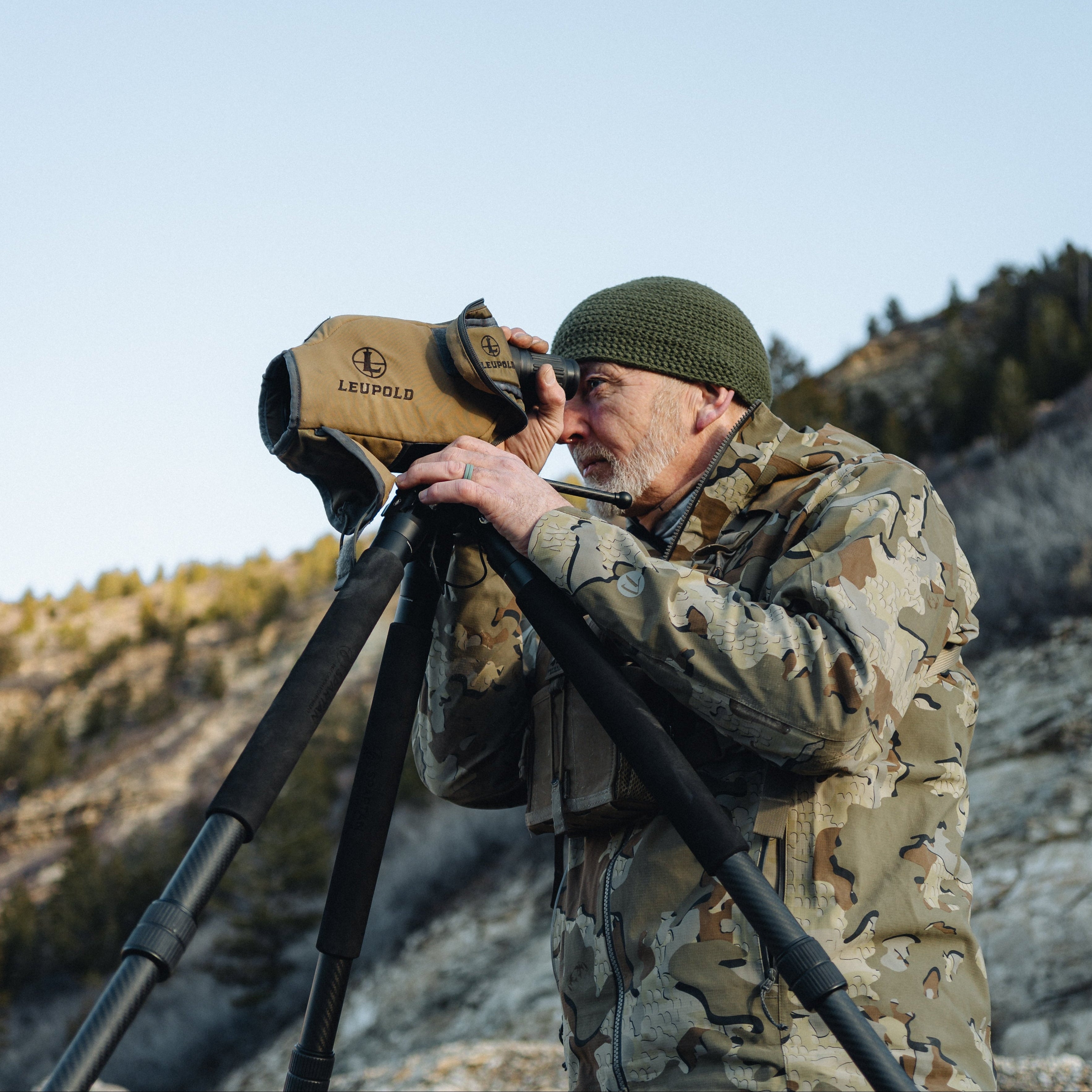 Hunter in camouflage gear using a Leupold spotting scope in a mountainous landscape.